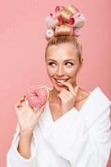 Portrait of pleased young housewife in housecoat with hair curlers smiling while eating sweet donut