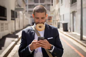Young professional man eating a doughnut and using smartphone on the street