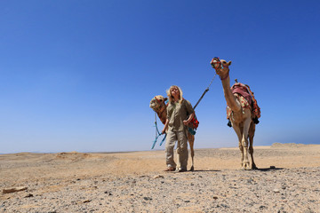 Wüstentrip: eine blonde Frau in outdoor Kleidung führt zwei Kamele durch die Wüste
