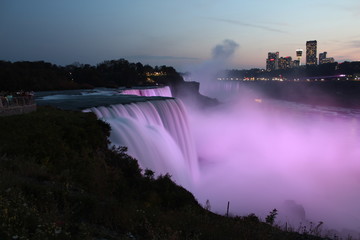 Wide view of Niagara Falls illuminated pink in evening as seen from USA, with water flow all blurred, cityscape visible on Canada side