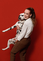Cute happy woman in white shirt and pants, is holding dalmatian dog in her arms on red background. Love to the animals, pets friendship concept. Copy space