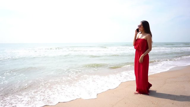 Static shot of a beautiful sexy lady wearing a red beach dress standing in front of the sea waves while talking on her phone