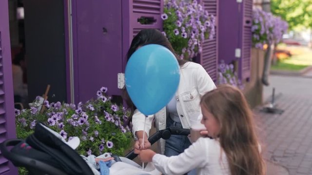 Young mother and her daughter playing with a baby in a stroller using a blue balloon while laughing and smiling
