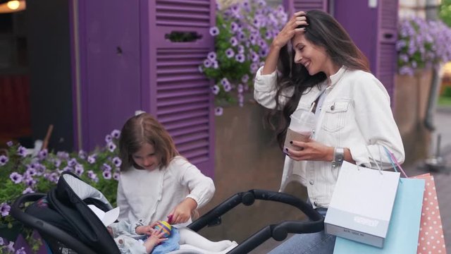 Medium handheld shot of beautiful young mother with her daughter playing with a baby bow sitting in a stroller and drinking smoothies