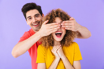 Photo of cheerful couple, man smiling and covering woman's eyes standing behind her