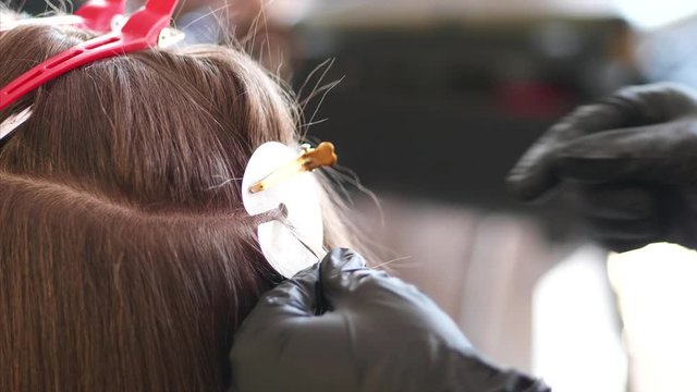 A hairdresser with two hands in black gloves is slowly installing the upgrade of hair extension for the woman client at the hair studio. Action of extension is close up.