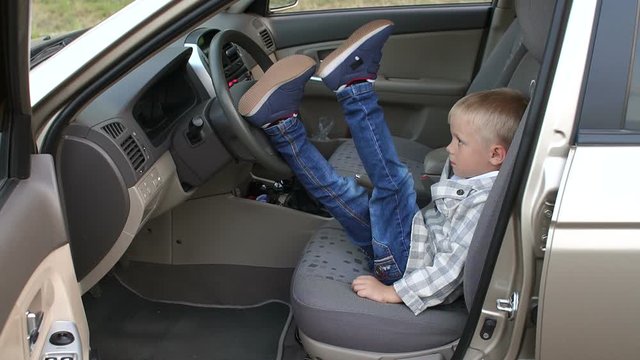 A small business boy sitting behind the wheel of a car in a jacket and jeans, he threw his feet on the steering wheel. Slow motion.