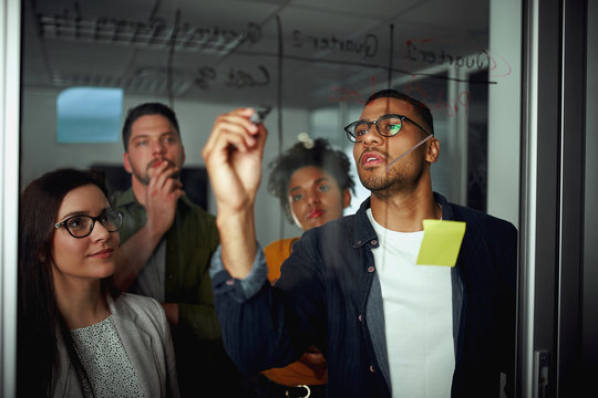 Pensive Diverse Group Of Young People Looking At Businessman Writing Business Future Plans On Glass During Office Meeting