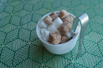White deep glass bowl with white and dark pieces of sugar. Tweezers also sharpen in a bowl. A bowl is standing on a green tablecloth