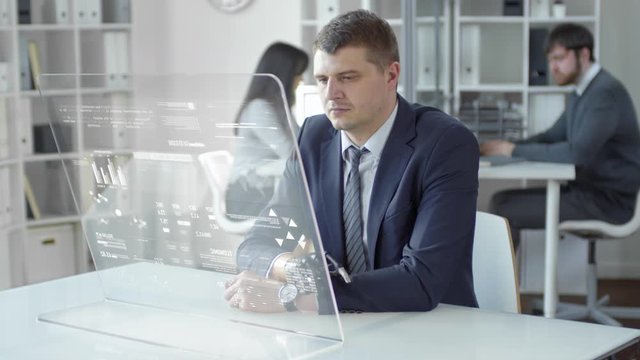 Businessman with prosthetic hand sitting at desk in the office and using computer with augmented reality screen while his colleagues working in the background