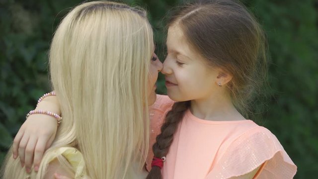 Portrait of mom kissing her little daughter. Cute blond mother and her little daughter hugging in the summer park. Happy loving family. Woman and girl together outdoors.