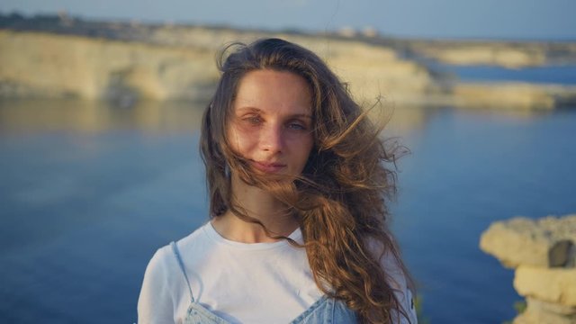 A young brunette woman is standing on a cliff, her hair is developing in the wind. Summer evening. Close-up 