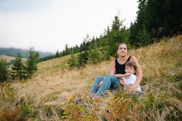 Naklejka na ściany i meble Smiling female hugs her son resting as they walking in mountains together