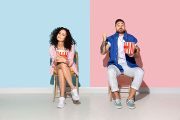 Fototapeta na wymiar Young emotional caucasian couple in bright casual clothes posing on pink and blue background. Concept of human emotions, facial expession, relations, ad. Man and woman watch cinema with popcorn.
