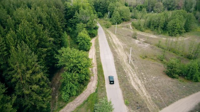 Aerial drone view following black modern van vehicle on countryside slab road between green coniferous pine trees. Car driving by dirt country way along forest. Tourism and travel.