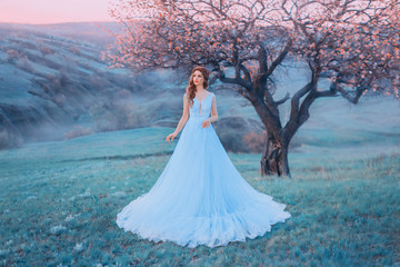incredible woman stands in the background of a mountain landscape in a blue dress. Tale of Cinderella with a doll face. Cold color. Delicate pink sky and a blossoming tree on a hill. Art photography