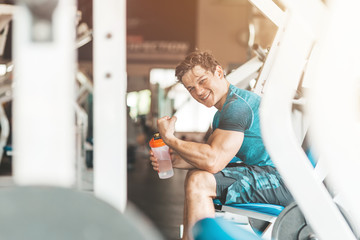 Portait of young man looking at camera while sitting in gym and holding a classic fitness shaker with pre-workout drink in it. Sports nutrition concept. Horizontal shot. Side view