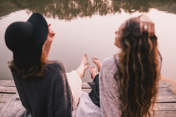 Two female friends in knitted warm sweaters having picnic near lake with autumn forest and lake on the background. Cozy fall atmosphere. 