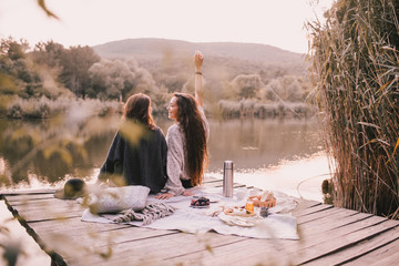 Two female friends in knitted warm sweaters having picnic near lake with autumn forest and lake on the background. Cozy fall atmosphere. 