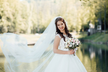 Fototapeta na wymiar Bride with bouquet near lake. Beautifully fluttering veil. Beautiful brunette bride makeup and hairstyle in luxury dress. The bride is holding a wedding bouquet of unusual beautiful flower. 
