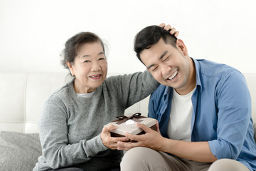 Happy Asian man giving gift box to his mother for Mother's Day celebration.