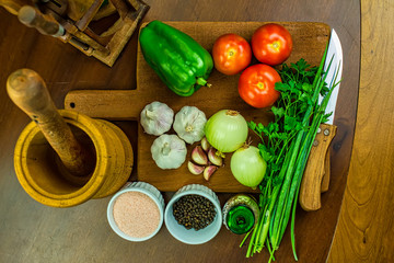 Garlic, onions, green peppers, tomatoes and parsley and chives on wooden cutting board with glass of olive oil, salt and black pepper in ramekins, wooden mortar and wooden chopping block in top view