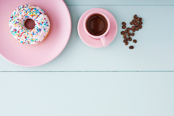 One white donut or doughnut in a pink plate, cappuccino in a pink coffee cup and coffee beans on a pastel blue wooden table with copy space. Top view.