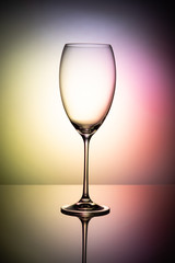 Empty glass goblet without wine on a thin leg stands on a mirror surface. Colorful background.