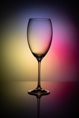 Empty glass goblet without wine on a thin leg stands on a mirror surface. Colorful background.
