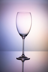 Empty glass goblet without wine on a thin leg stands on a mirror surface. Colorful purple background.