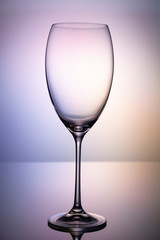 Empty glass goblet without wine on a thin leg stands on a mirror surface. Colorful purple background.