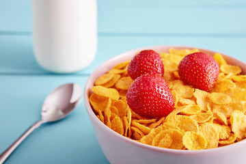 Bowl of dry corn flakes cereal with strawberry topping, a bottle of milk and a spoon on blue wooden country style table. Close up view.
