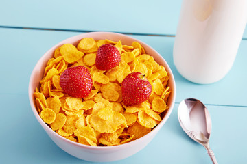Bowl of dry corn flakes cereal with strawberry topping, a bottle of milk and a spoon on blue wooden country style table. Close up view.