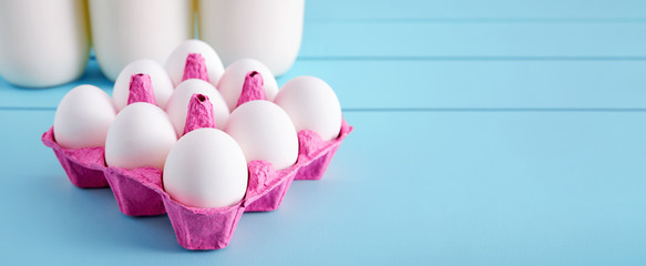 White eggs in a pink filler and milk bottles on turquoise wooden country kitchen table. Banner with copy space