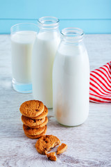 Two bottles of milk, chocolate chip cookies and a glass full of milk on wooden table with blue wooden background.