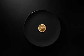 One dry lemon slice served in black plate on moody black background. Top view.