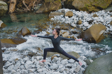 Young slim woman practicing yoga outdoors on stone shore of mountain river. Unity with nature concept. Girl standing in warrior pose