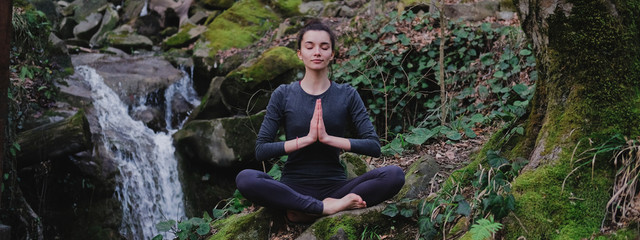 Young slim woman practicing yoga outdoors in moss forest on background of waterfall. Unity with nature concept. Girl meditates sitting