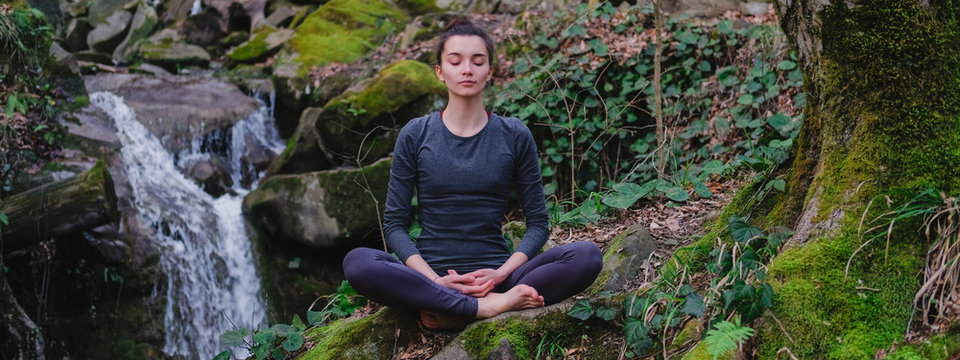 Young slim woman practicing yoga outdoors in moss forest on background of waterfall. Unity with nature concept. Girl meditates sitting