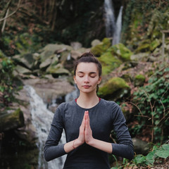 Young slim woman practicing yoga outdoors in moss forest on background of waterfall. Unity with nature concept. Girl standing with prayer hands