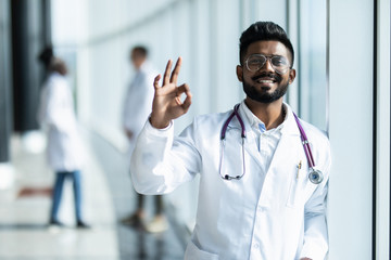 Young indian male doctor with okay sign in white uniform with collegues on the background