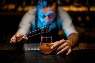 Male bartender adding chilled melting caramel with twezzers to the cocktail with ice cubes under blue light