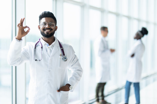 Young Indian Male Doctor With Okay Sign In White Uniform With Collegues On The Background