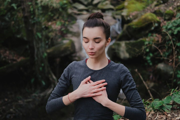 Young woman practicing breathing yoga pranayama outdoors in moss forest on background of waterfall....
