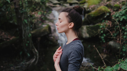 Young woman practicing breathing yoga pranayama outdoors in moss forest on background of waterfall....
