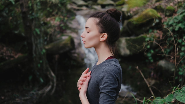 Young woman practicing breathing yoga pranayama outdoors in moss forest on background of waterfall. Unity with nature concept.