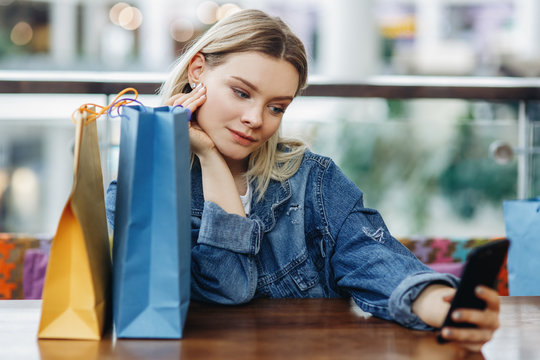 Portrait of beautiful blonde woman in a jeans jacket with shopping bags sitting in cafe at shopping mall. She is taking pictures of herself on her phone doing a sephi. Copy space on the right side