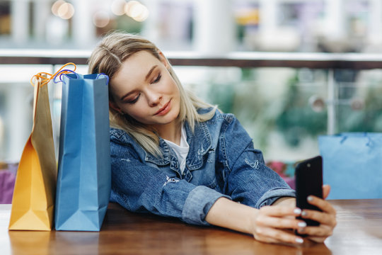 Portrait of woman in a jeans jacket with shopping bags sitting in cafe at shopping mall. She is taking pictures of herself on her phone doing a sephi. Copy space on the right side