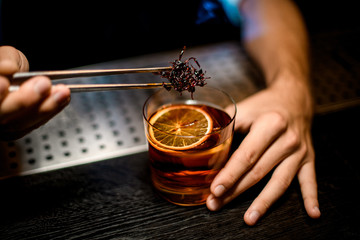 Male bartender adding chilled brown caramel with twezzers to the cocktail with ice cubes and dried orange slice
