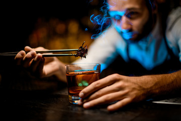 Bartender adding chilled melting caramel with twezzers to the cocktail with dried orange under blue light